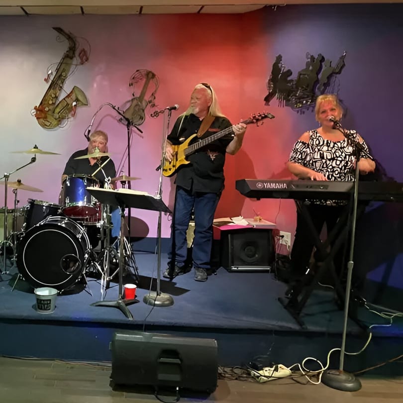 A three-person band performs on a stage with musical instrument wall decor at Galindo's.