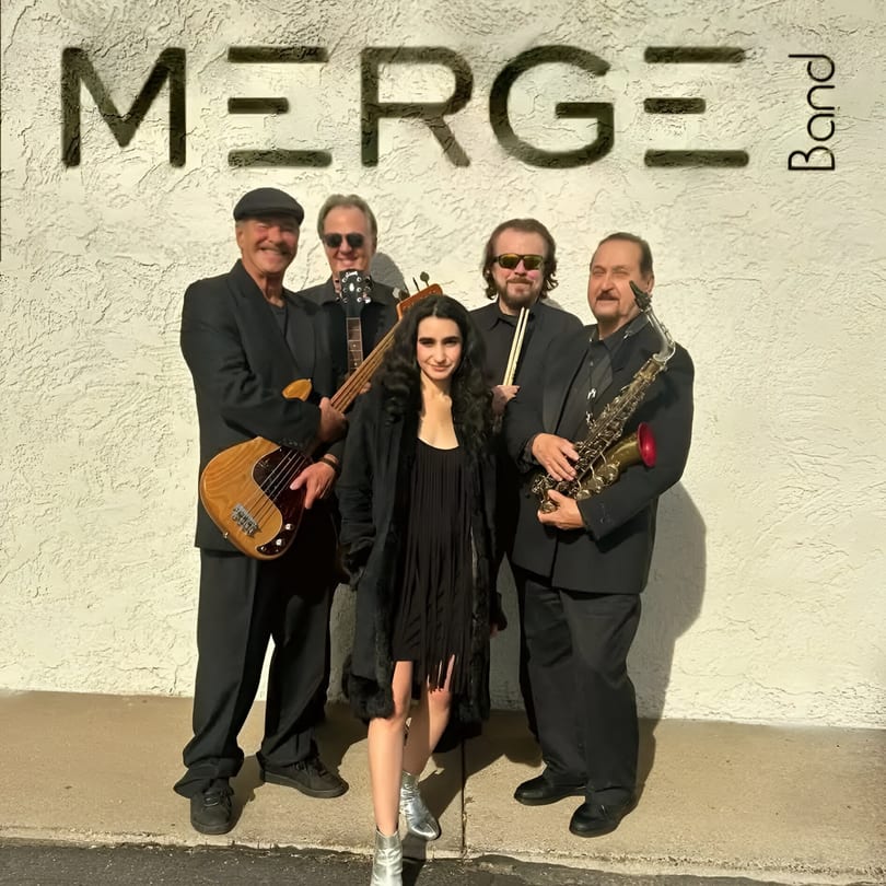 Five band members, four men and one woman, posing with instruments in front of a white wall.