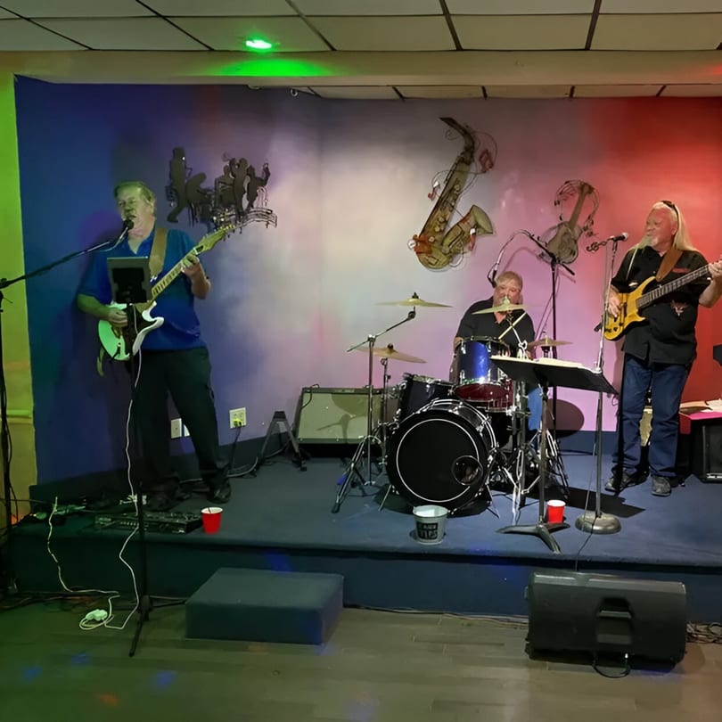 A three-person band performs on a stage with colorful lighting and musical instrument wall art.
